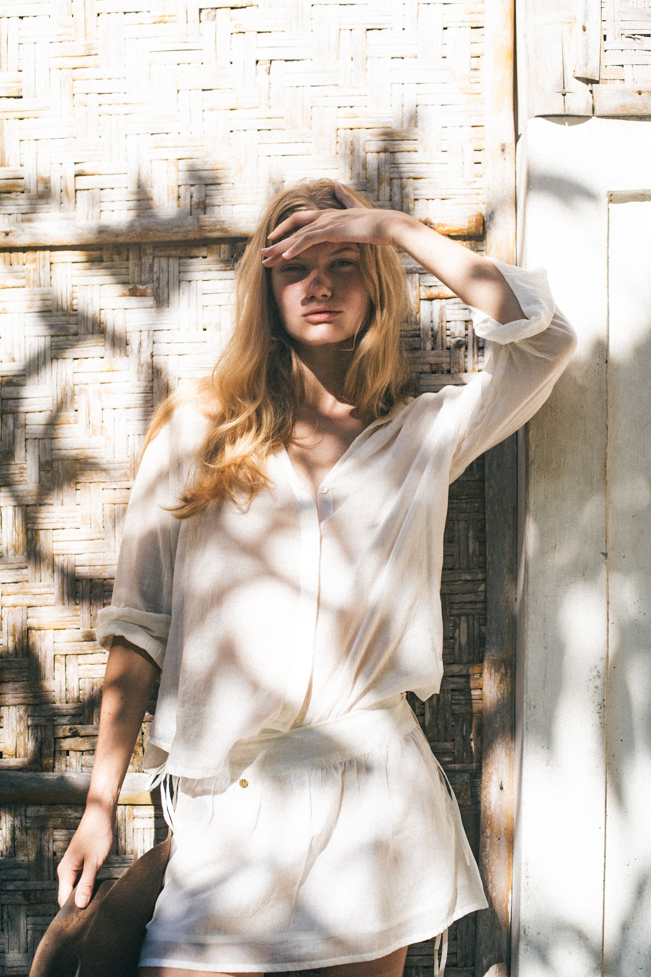 Woman in a white shirt standing against a textured wall with sunlight casting shadows.